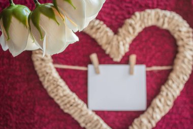 Beyaz gül tomurcukları, kalp şeklinde örülmüş bir çerçeveye karşı kırmızı zemin üzerinde bir kartla.