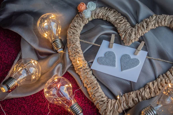 Плетеная рамка в форме сердца с карточкой на спицах на серой ткани рядом с лампочками — стоковое фото
