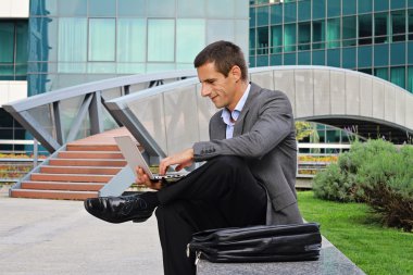 Young handsome businessman, manager using laptop outdoors in the city, in front of modern building.