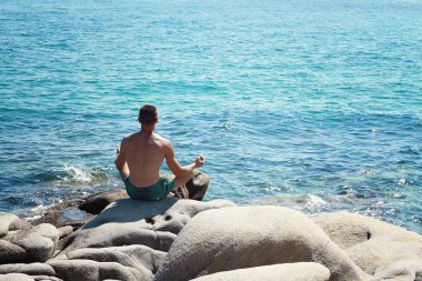 Man Meditating Near The Sea sitting on a rock and Doing Yoga On the Beach in a lotus position