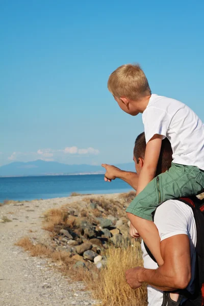 Father giving his son piggyback ride outdoors. Man and boy son infront of sea, pointing away, active summer holiday vacation, family travel photo