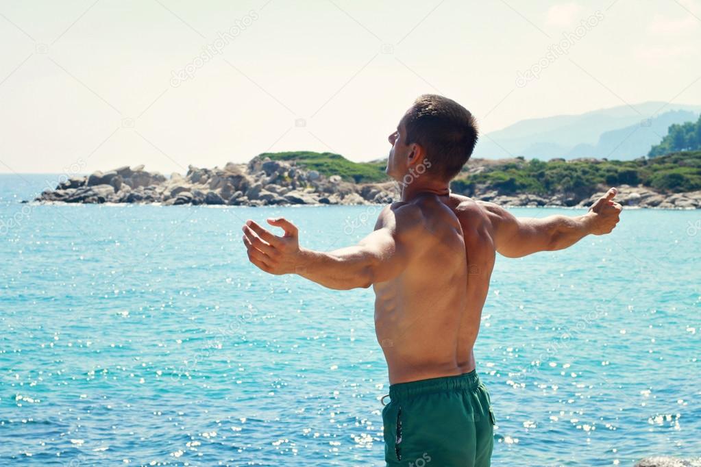 Man Raising His Hands or Open arms  looking to sea blue sky horizon. Strong muscular Men, perfect body, arms, back. Freedom Concept