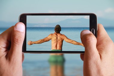 Male hand taking photo of Man Raising His Hands or Open arms standing back looking to sea blue sky horizon with cell, mobile phone. Strong muscular Men, perfect body, arms, back. Freedom Concept