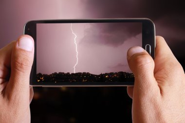Male hand taking photo of lightning in the city lanscape with cell, mobile phone.