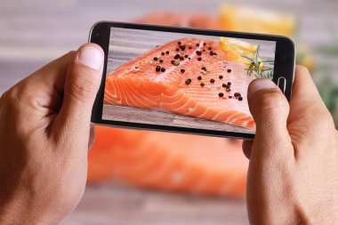 Male hand taking photo of Raw salmon fillet with lemon and herbs on wooden background with cell, mobile phone.