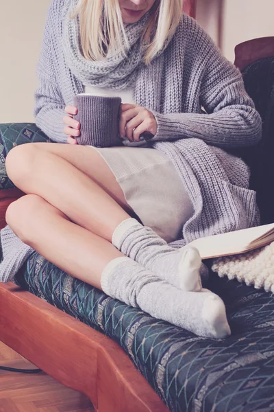 Beautiful Woman wearing  cozy sweater and Warm Wool Socks  reading a book sitting on comfortable sofa enjoying hot chocolate drink . Winter, Christmas  holidays concept.