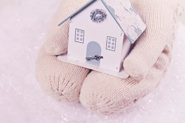 Female hands in white knitted cozy mittens holding cute house on a white snow background. Christmas tree decoration. Family, Home, Love concept. Winter and Christmas time — Stock Photo, Image