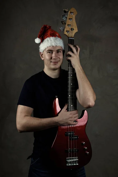 Молодой человек в новогодней кепке с гитарой. Праздник. Серый фон — стоковое фото