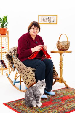 Büyükanne bir sandalyeye oturur ve örgü örer, gri bir kedi yanına oturur.