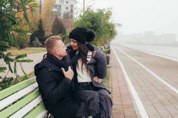 Mladý zamilovaný pár sedí na lavičce na náspu, usmívá se a dívá se jeden druhému do očí — Stock fotografie