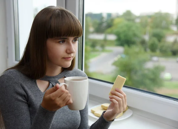 The girl is sitting by the window with a cup of tea in her hands.