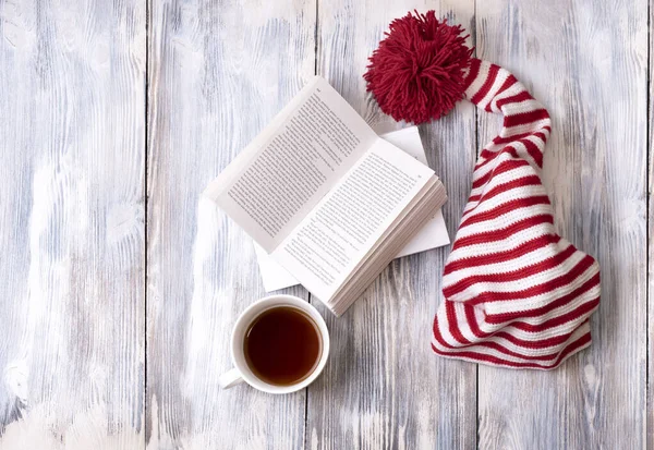 Büyük kırmızı ponponu olan kırmızı ve beyaz çizgili bir şapka ahşap beyaz bir zemin üzerinde kitap ve bir fincan çay, kopya alanı, üst görünüm.