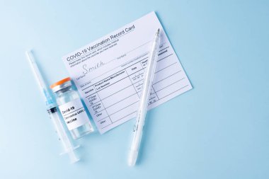 Covid-19 vaccine vial, vaccination record card, syringe, a pen on a blue background, copy space, close up. Coronavirus vaccination concept.