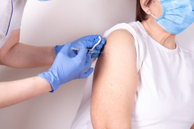 Gloved hands injectng a syringe with covid-19 vaccine into the shoulder of an old woman, close up.