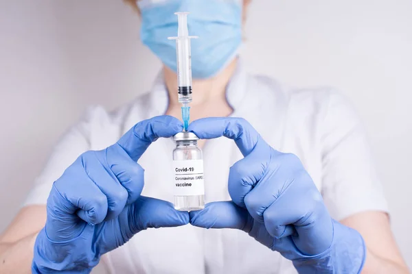 A woman doctor in a mask and gloves holding a vial with covid-19 vaccine and syringe on a white background, close up. Coronavirus vaccination.