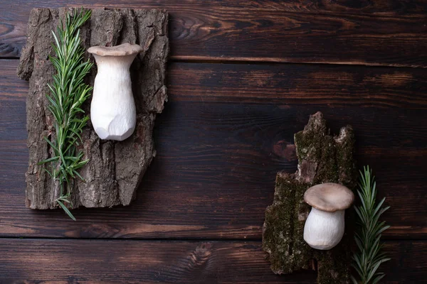 Two fresh king trumpet mushrooms with a sprigs of rosemary on a bark on a wooden background, rustic style, close up.