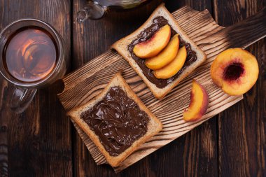 Çukulata ezmeli tost, şeftali dilimli bir tabak, bir fincan çay, koyu ahşap bir arka planda kesim tahtası, tatlı atıştırmalık..