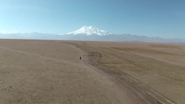 Mulher menina em keds brancos t-shirt denim macacões andando em terras altas no pico Elbrus nevado. Bela paisagem de drone voando sobre o campo Elbrus ápice. Paisagem montanhosa Área de Kabardino-Balkaria — Vídeo de Stock