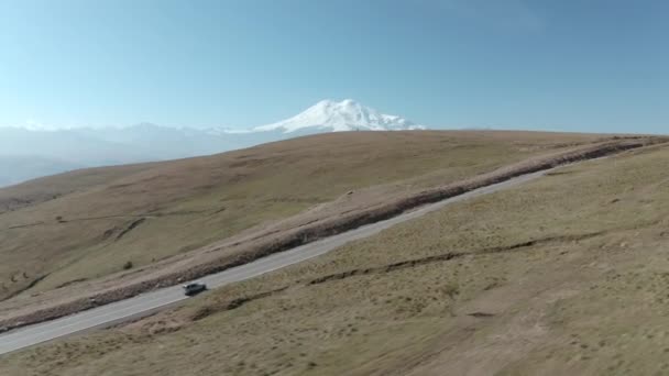 Ταξιδιωτικό αυτοκίνητο οδήγηση σε ορεινό δρόμο σε χιονισμένο ορεινό τοπίο κορυφή. Αυτοκίνητο SUV κινείται στην εθνική οδό προς Dzhily Su σε Kabardino-Balkaria. Αεροπλάνα πλευρά από ιπτάμενο drone — Αρχείο Βίντεο