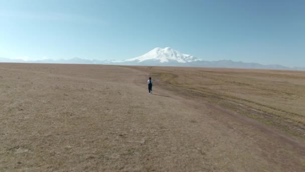 Mulher correndo no campo na neve Elbrus pico paisagem. Vista aérea menina camiseta branca macacão jeans no campo highland em Elbrus panorama pico nevado. Bela paisagem montanhosa Kabardino-Balkaria — Vídeo de Stock