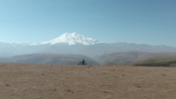 自転車の女性は雪の山のピークの背景に自転車に乗る。スポーツエンデューロの旅の観光の女の子雪の上の山の平野上の完全なサスペンションe-bike自転車エルブラスの風景 — ストック動画