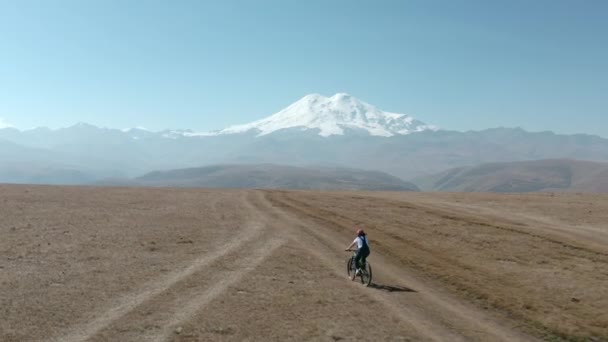 雪上の山の平野やドローンからの谷の風景で電動自転車に乗る女性。高地雪エルブラスDzhily蘇航空風景のマウンテンバイクでの旅行者の女性の自転車 — ストック動画