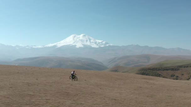 Sport nő lovaglás elektromos kerékpár sivatagi területen, míg a kerékpár utazás havas Elbrus. Utazó lány lovaglás e-bike hegyi terepen havas csúcs légi táj — Stock videók