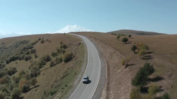 SUV coche gris que conduce en la carretera de las tierras altas en el paisaje nevado pico de montaña. Automóvil moviéndose en asfalto camino vacío a Dzhily Su en Kabardino-Balkaria. Paisaje aéreo de las tierras altas desde el dron arriba — Vídeos de Stock