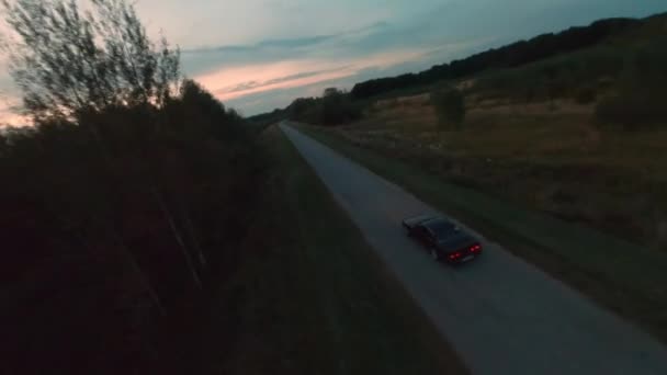 Czarny samochód jdm jazdy po wiejskiej drodze na tle wieczornego zachodu słońca. Fpv drone kinematograficzny widok sport pojazdu jazdy w zmierzchu na autostradzie podmiejskiej na drzewach letnich i krajobraz horyzontu — Wideo stockowe