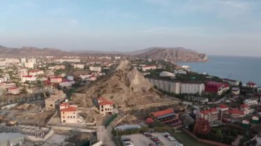 Karadeniz kıyısındaki Sudak şehrindeki insansız hava aracının panoramik görüntüsü. Gökyüzü manzarası dağ kayaları ve ufuktaki mavi deniz kıyısındaki modern şehir merkezi