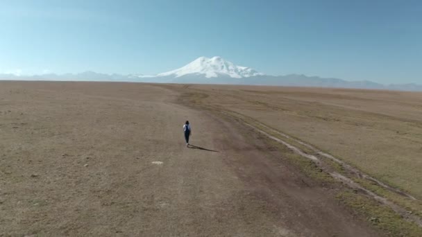 Női lány fehér keds t ing farmer overál megy előre a hegyekbe havas Elbrus csúcs. Légi táj a drónrepüléstől az Elbrus-csúcs felett. Kabardino-Balkaria hegyvidék. — Stock videók