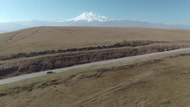 Vehículo de viaje conduciendo en carretera asfaltada de tierras altas en el paisaje nevado pico de la montaña. SUV coche que se mueve en la carretera a Dzhily Su en Kabardino-Balkaria. Disparo lateral aéreo desde un avión no tripulado volador — Vídeo de stock