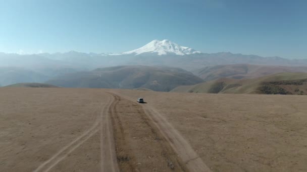 Fantastisk antenn framåt vy från höglandet till snöig topp Elbrus topp. Resor SUV bil stående på off road fält med panoramautsikt snöig topp Elbrus fäste i Kabardino-Balkaria, Dzhily Su område — Stockvideo