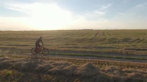 Motociclista en paseos casco verde mtb electro suspensión completa bicicleta de montaña e-bike a lo largo de polvo de carretera sucia rural de las ruedas más allá del campo y la madera en la soleada puesta de sol de otoño por la noche aérea alrededor de la vista — Vídeos de Stock