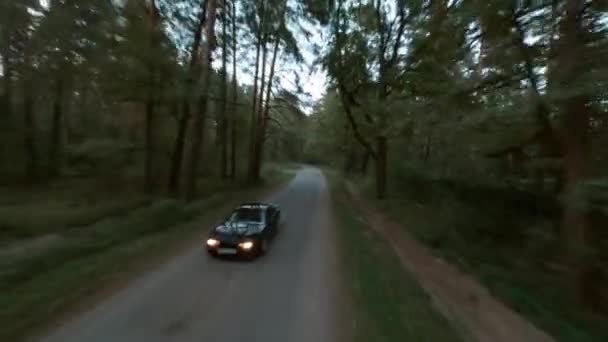 Voiture sport noire avec phares sur les vitesses le long de la route rurale étroite vide à travers la vieille forêt de pins en automne en fin de soirée première vue aérienne cinématographique prise de vue de drone de course fpv — Video