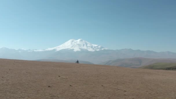 Person rider mtb cykel längs landsvägen i fält mot avlägsna Elbrus berg under klar himmel på solig dag antenn fågel ögon — Stockvideo