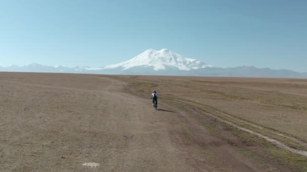 在蓝天蓝天的天空下，妇女游客骑着轻便摩托车沿着空旷的乡间道路在田野里与白雪覆盖的Elbrus山相对照 — 图库视频影像