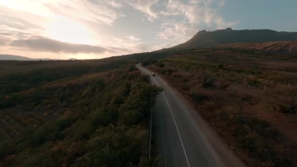 Czarny samochód cruising jazdy wzdłuż pustej asfaltowej autostrady biegnącej przez Highland Mountain Valley pod zachmurzonym słońcem niebo jesienią zachód słońca wieczorem fpv sport drone pierwszy punkt antenowy widok kinowy — Wideo stockowe