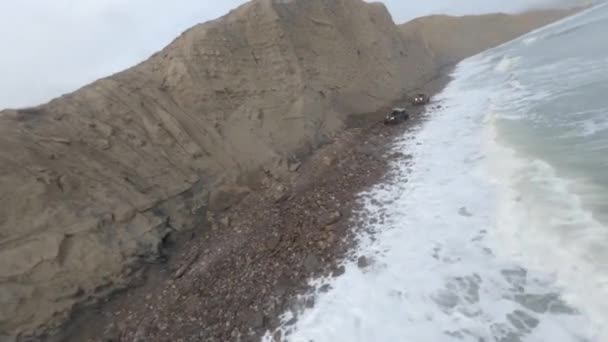 Aerial shot flying over two rally cars riding on pebble waving sea beach coast with mountains — Stockvideo