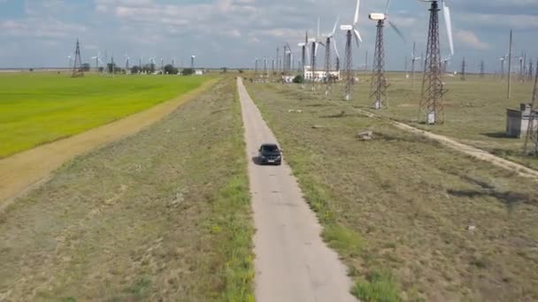 Carro preto dirigindo no asfalto estrada moinho de vento turbina fundo no campo agrícola verde. Drone vista aérea lateral estação de energia eólica. Turbinas eólicas geradoras de energia renovável limpa — Vídeo de Stock