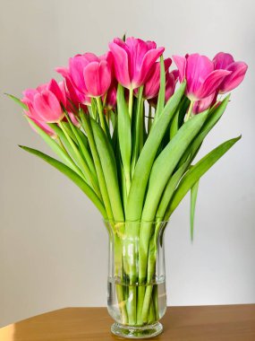 Gri arka planda bir masanın üzerinde pembe laleler olan şeffaf bir vazo.