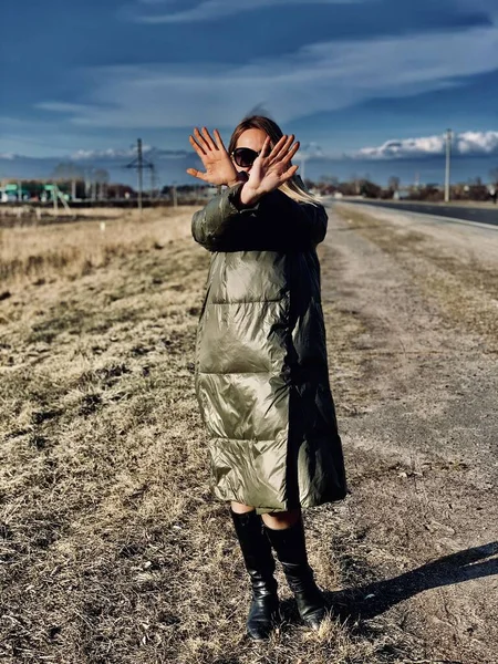 young woman in a green jacket, coat. Young woman covers her face with her hands. young woman stands against a background of clouds.