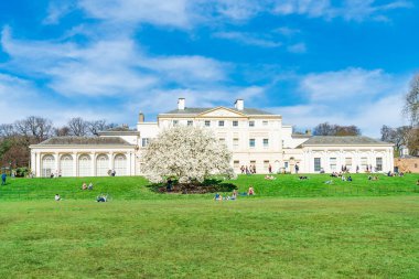 LONDON, UK - MARCH 30, 2021: People enjoy sunshine outside Kenwood House with beautiful white magnolia tree blooming in a the park on the northern boundary of Hampstead Heath clipart