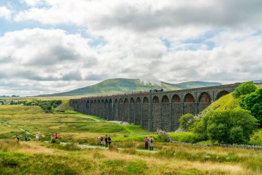 RIBBLEHEAD, UK - AUGUST 24, 2021: Train spotters watch a steam train passing over the Grade II listed Ribblehead Viaduct in the Ribble Valley at Ribblehead in North Yorkshire, England clipart