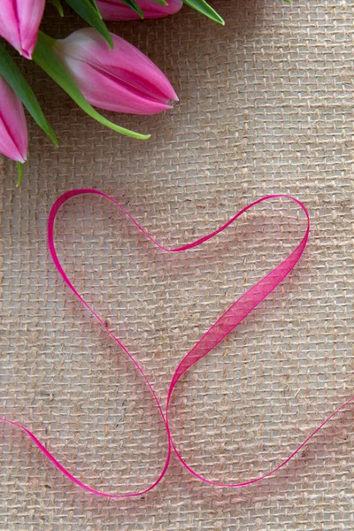 Cinta roja del corazón y tulipanes rosados aislados sobre fondo de tela marrón. —  Fotos de Stock