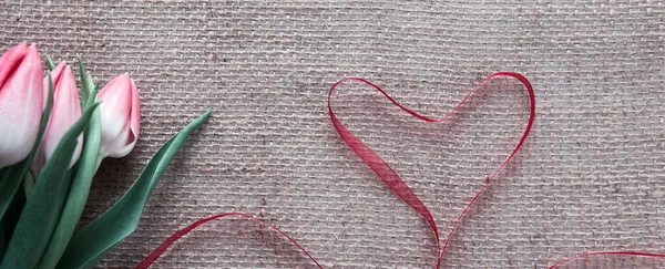 Cinta roja del corazón y tulipanes rosados aislados sobre fondo de tela marrón. — Foto de Stock