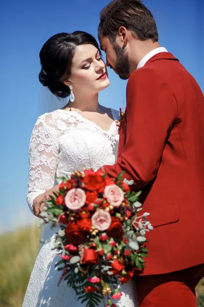 新郎和新娘，可爱的夫妻户外，婚礼新娘花束开红色花。蓝色的天空，绿草在背景中. — 图库照片