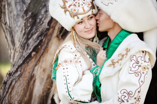 Coppia in costumi etnici abbracciano sullo sfondo di legno strutturato, sposo bacia sposa a guancia . — Foto Stock