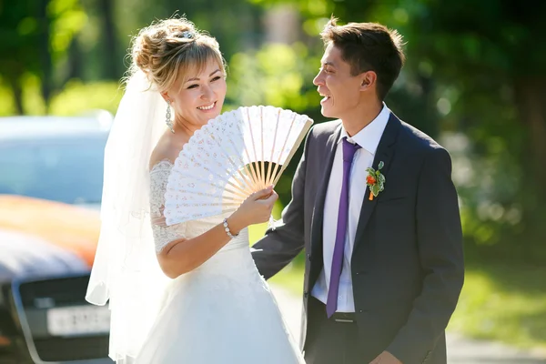 Noivo e noiva com ventilador na rua iluminado pelo sol, carro no fundo . Fotos De Bancos De Imagens