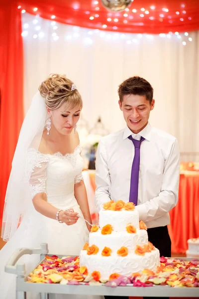 Braut und Bräutigam schneiden den Kuchen bei einem Bankett mit orangefarbenen Blumen zur Dekoration an. — Stockfoto